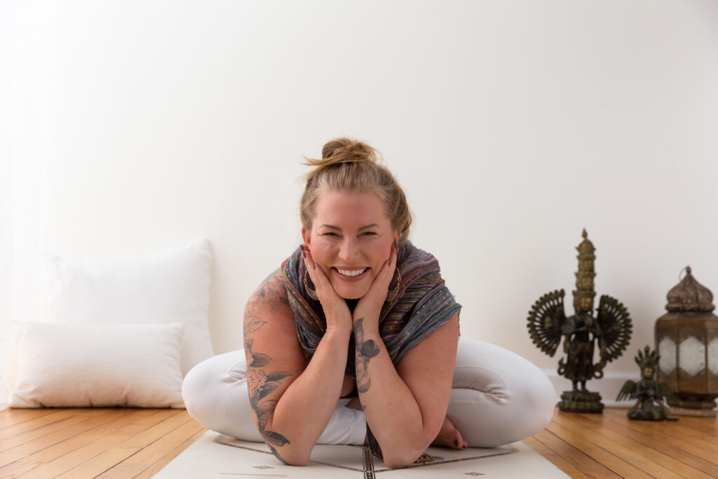 Brand image of yogi seated on floor, she hols her face in her palms with elbos leaning on floor, she is smiling and her tattoos show and her hair is in a messy bun atop her head.