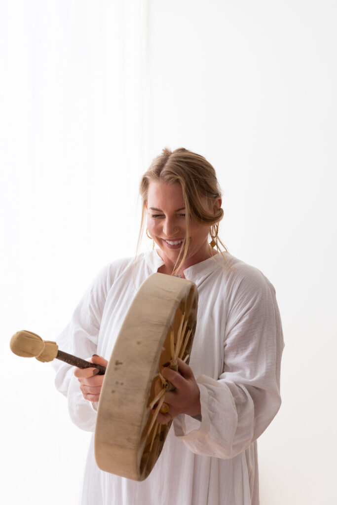 Brand image of same woman smiling and using a hand drum with a white bac ground wearing white tunic