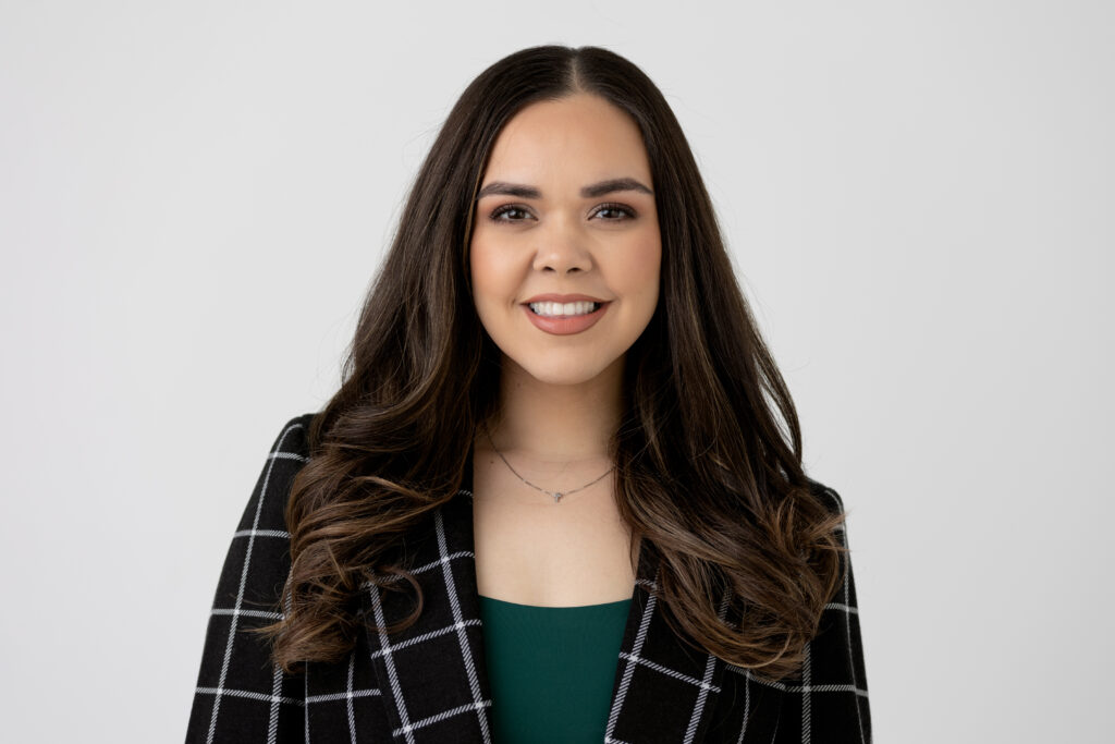 Professional Headshot of woman with long dark brown hair, black and white plaid blazer and green shirt, she is smiling in front of a white backdrop