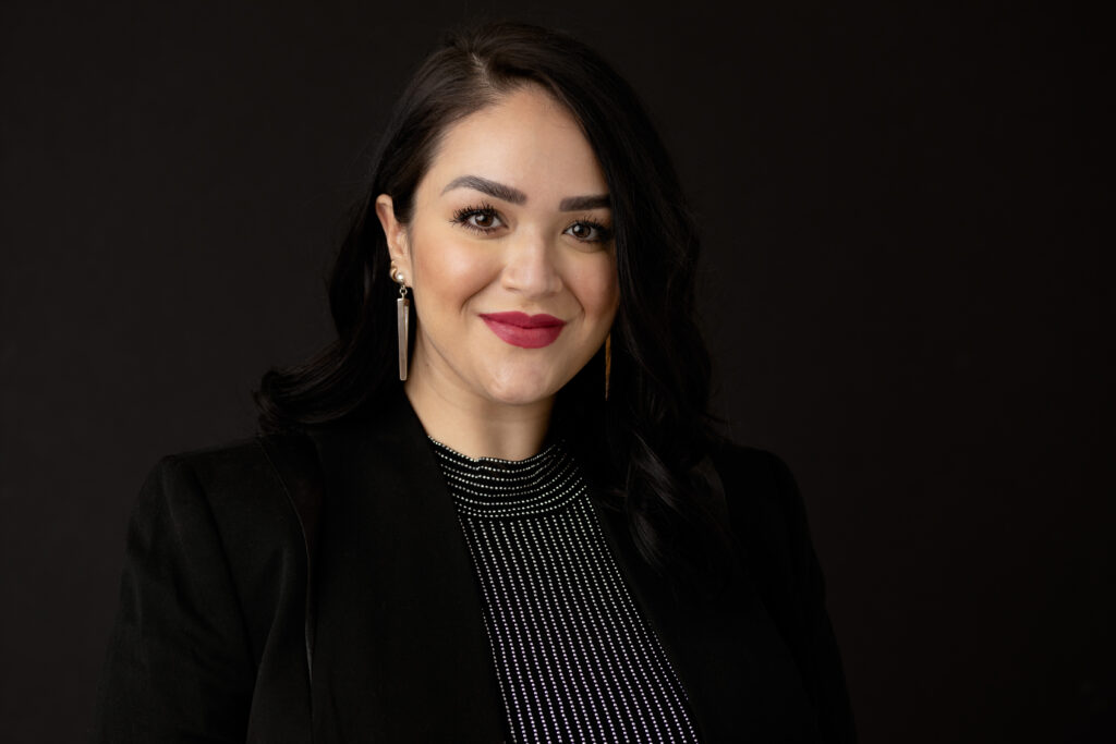 Professional headshot of women on a black backdrop, black blazer, dark grey top and bold red lipstick