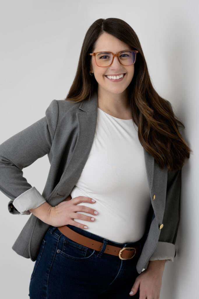 Professional headshot 3/4 length of young lady in glasses leaning on a wall with her hand on her hip