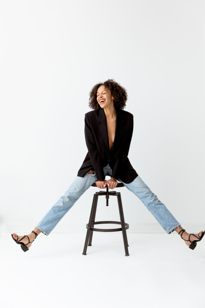 Young woman with natural curly hair balancing on a stool with legs splayed, she is excited, closed eyes and open mouth smile, she is wearing ripped straight leg jeans, black blazer with nothing under and strappy black sandles