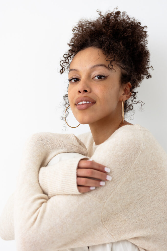 Woman in cream sweater embracing herself and looking at the camera with a soft smaile, her natural curly hair is piled on top of her head in a loose bun and she has tendrals falling around her face