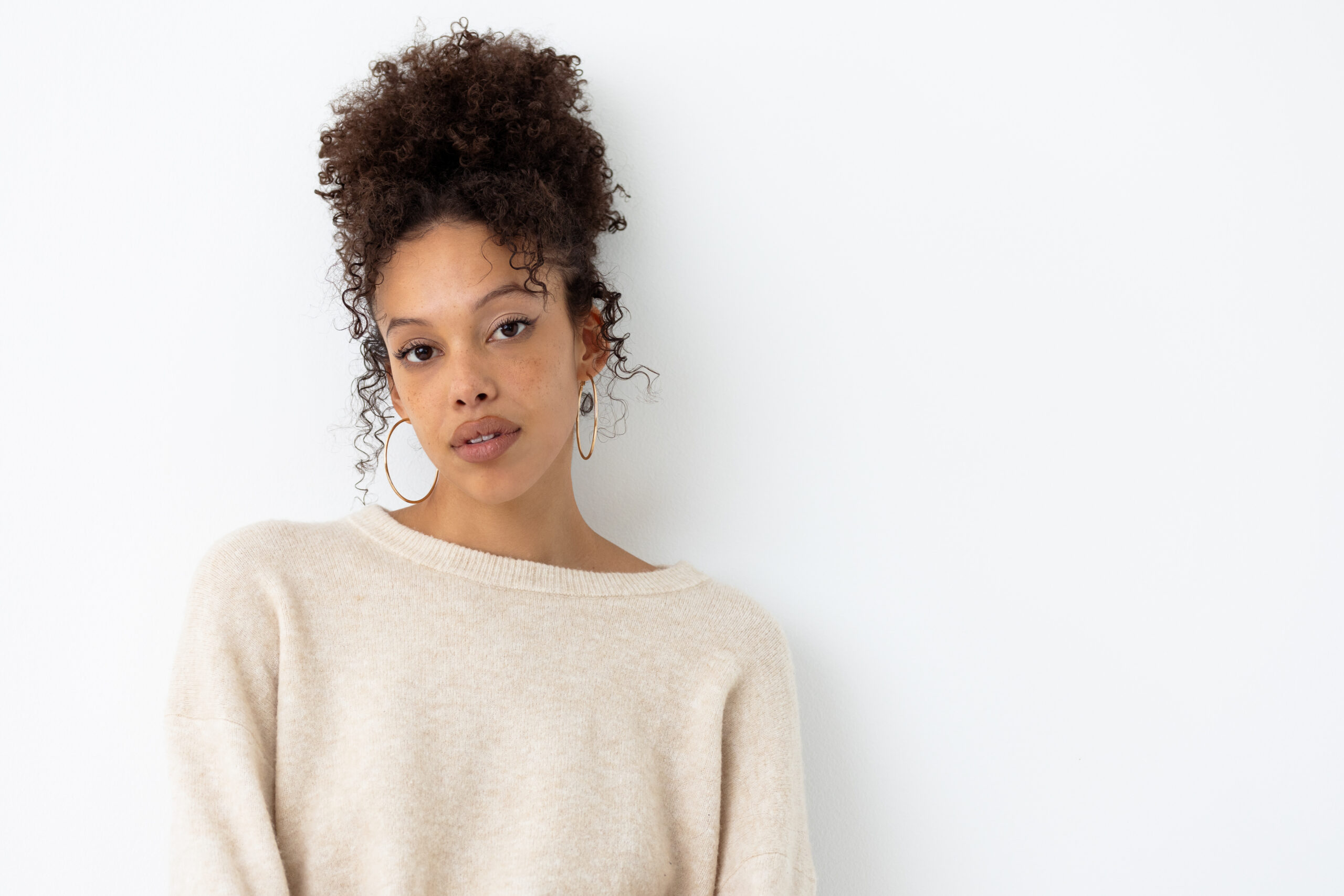 Woman with her natural curly hair in a loose bun on the top of her head, she is wearing a cream cashmere sweater and casually leaning back on a white wall.