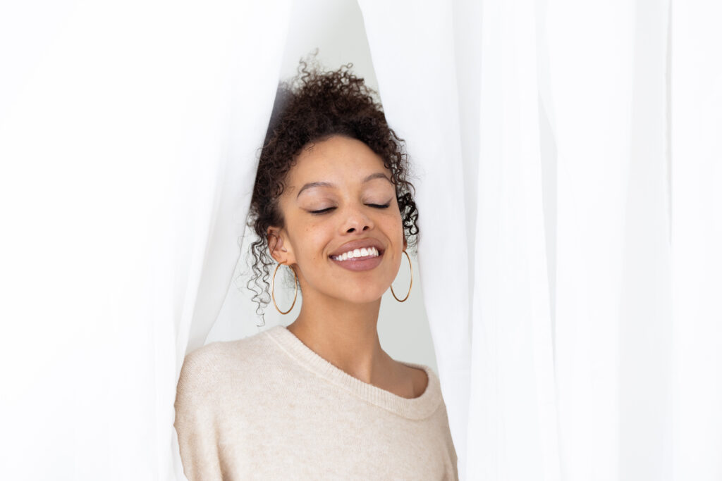 Women in a cream cashmere sweater, peaking through white curtains with a closed eye smile, enjoying herself