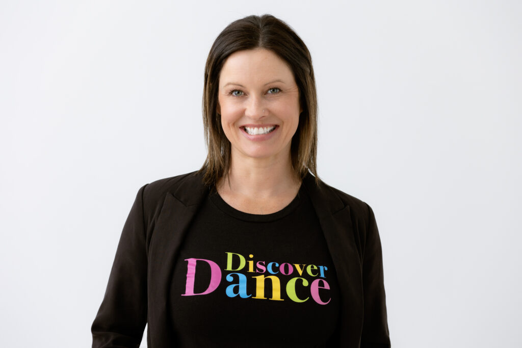Pro headshot with woman smiling on a white backdrop wearing a black blazer and a colorful branded black t-shirt