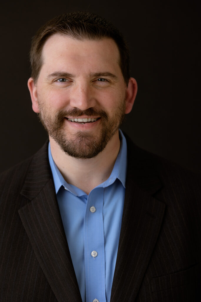 Professional headshot of man with facial hair, black blazer, blue collared shirt with a black background