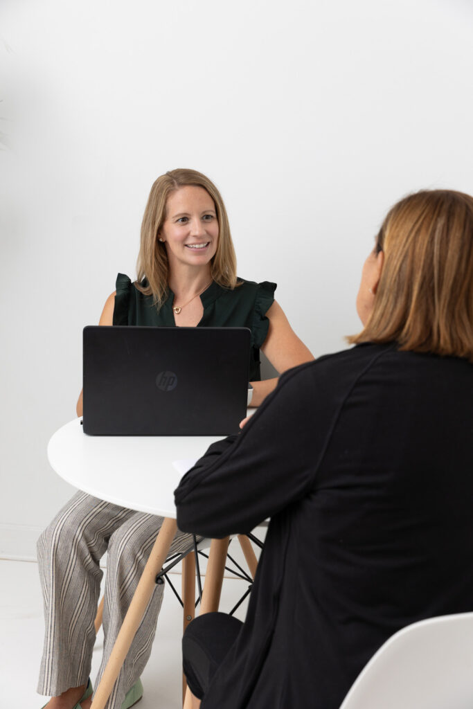 Two women are sitting at a small round table, engaged in conversation. One woman, smiling and facing the camera, has a laptop open in front of her. The other woman, seen from behind, listens attentively. The setting appears to be a casual meeting or interview.