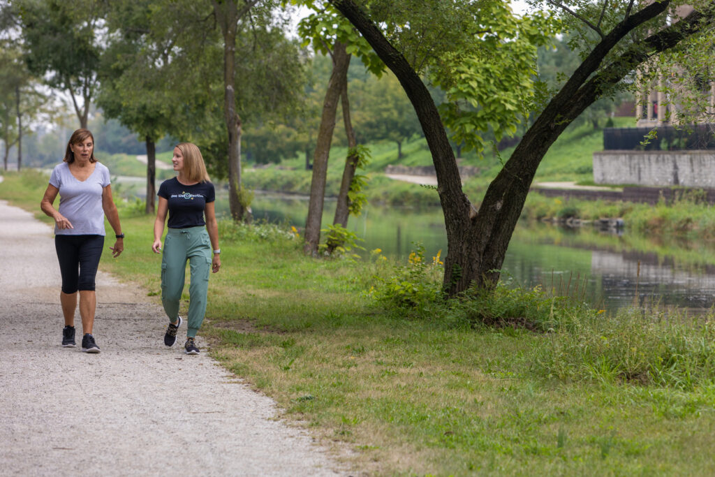 Two women are walking and talking on a gravel path next to a serene canal lined with trees.