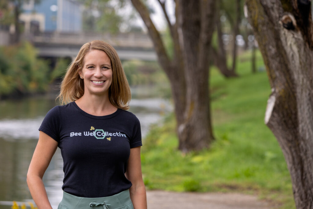 A woman stands smiling near a canal with trees and greenery in the background. She is wearing a black t-shirt with the words "Bee Well Collective" and small bee graphics, paired with green pants.