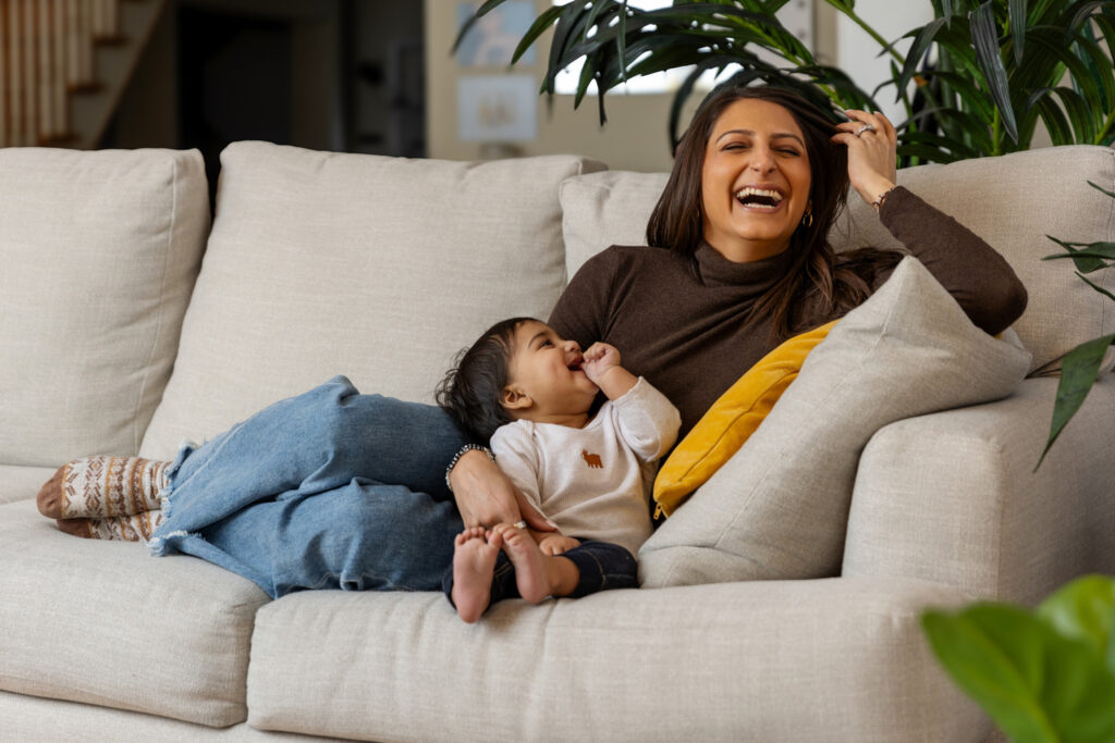 A woman is sitting on a beige sofa, laughing and holding a baby on her lap. She is wearing a brown turtleneck and blue jeans, while the baby is dressed in a white onesie with an orange animal design.