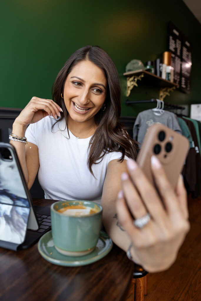 A woman is sitting in a café, smiling as she looks at her phone while holding it up for a selfie or video call. She is wearing a white t-shirt and bracelets, with a green coffee cup and a laptop in front of her.