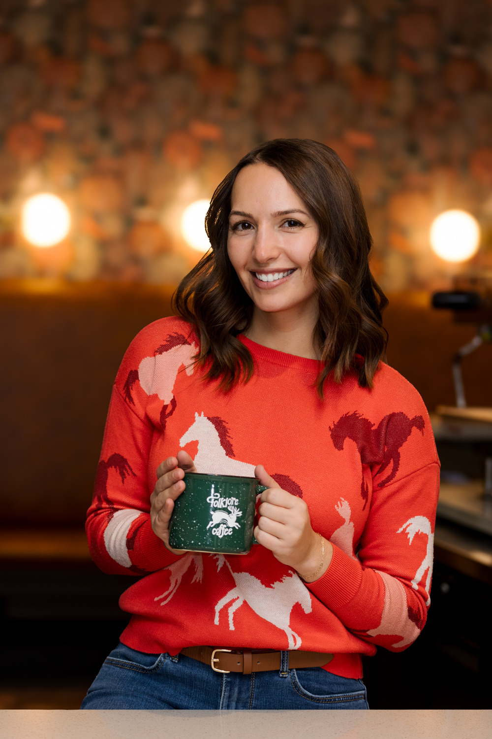 A woman smiles warmly at the camera while holding a green mug labeled "Folklore Coffee." She is wearing a bright orange sweater with a horse pattern, and the background features a softly lit, cozy coffee shop interior.