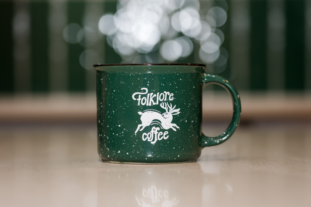 A green mug with a white "Folklore Coffee" logo, featuring a mythical creature, is placed on a reflective surface. The background is softly blurred with bokeh lights, creating a cozy and inviting atmosphere.