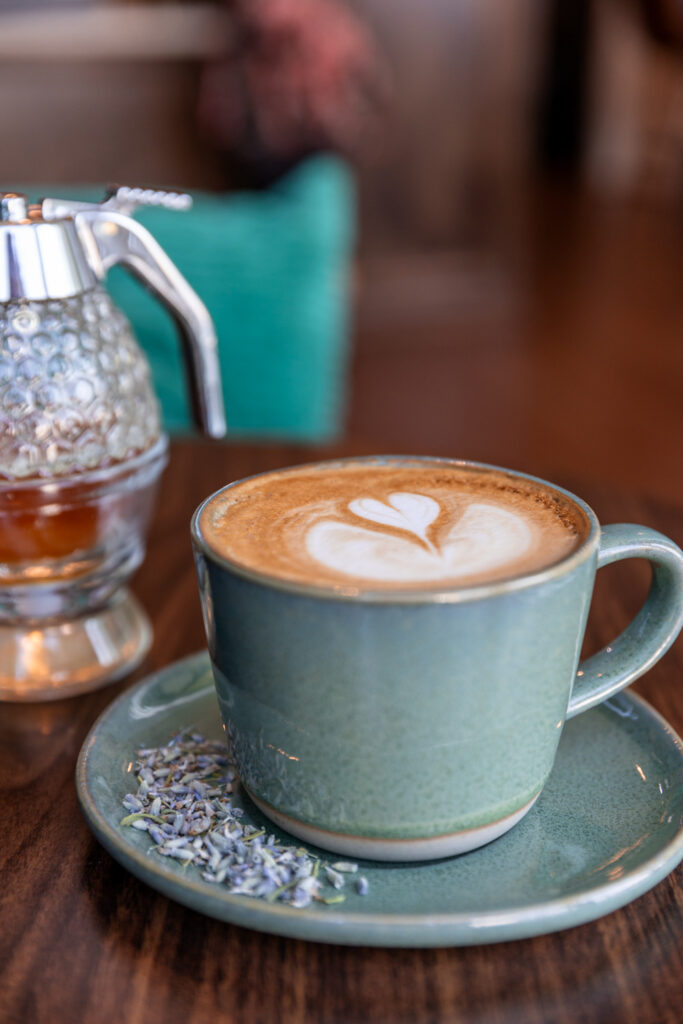 A green ceramic cup and saucer hold a latte with a heart-shaped latte art design, garnished with lavender petals on the saucer. Beside the cup is a vintage-style glass syrup dispenser. The background is softly blurred, highlighting the cozy and inviting ambiance of the coffee setting.