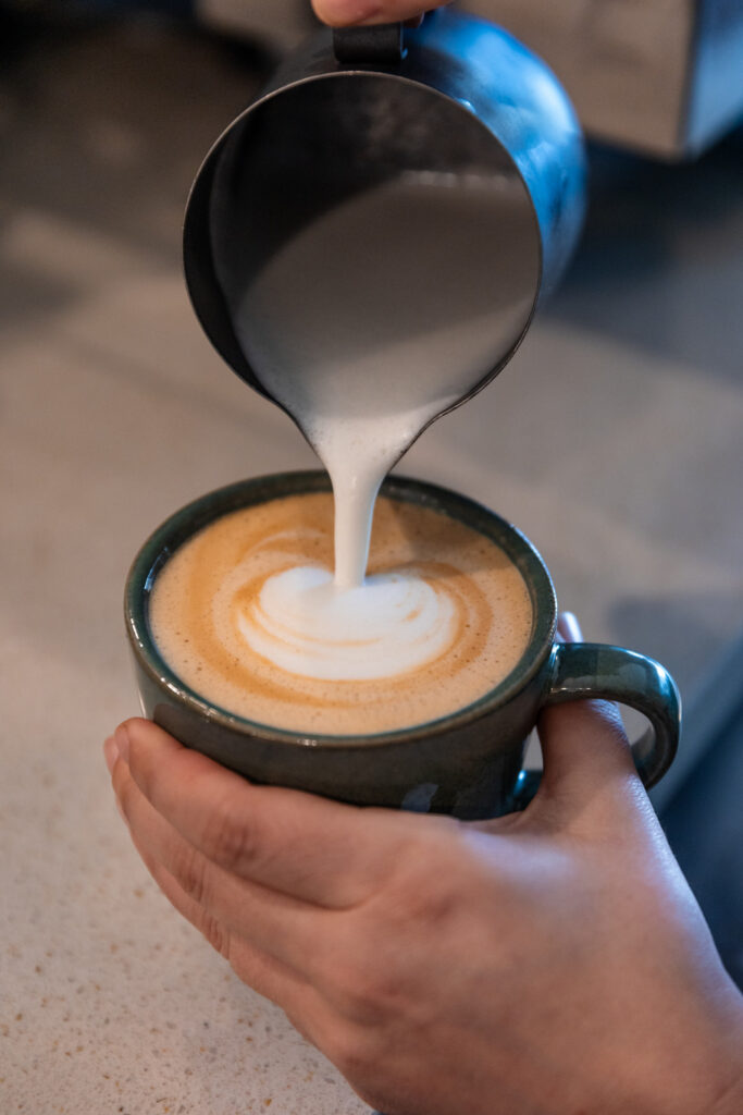 
A barista pours steamed milk from a metal pitcher into a cup of coffee, creating a latte art design. The close-up shot captures the skilled hands and the smooth flow of milk, emphasizing the precision and care in making the perfect latte. 