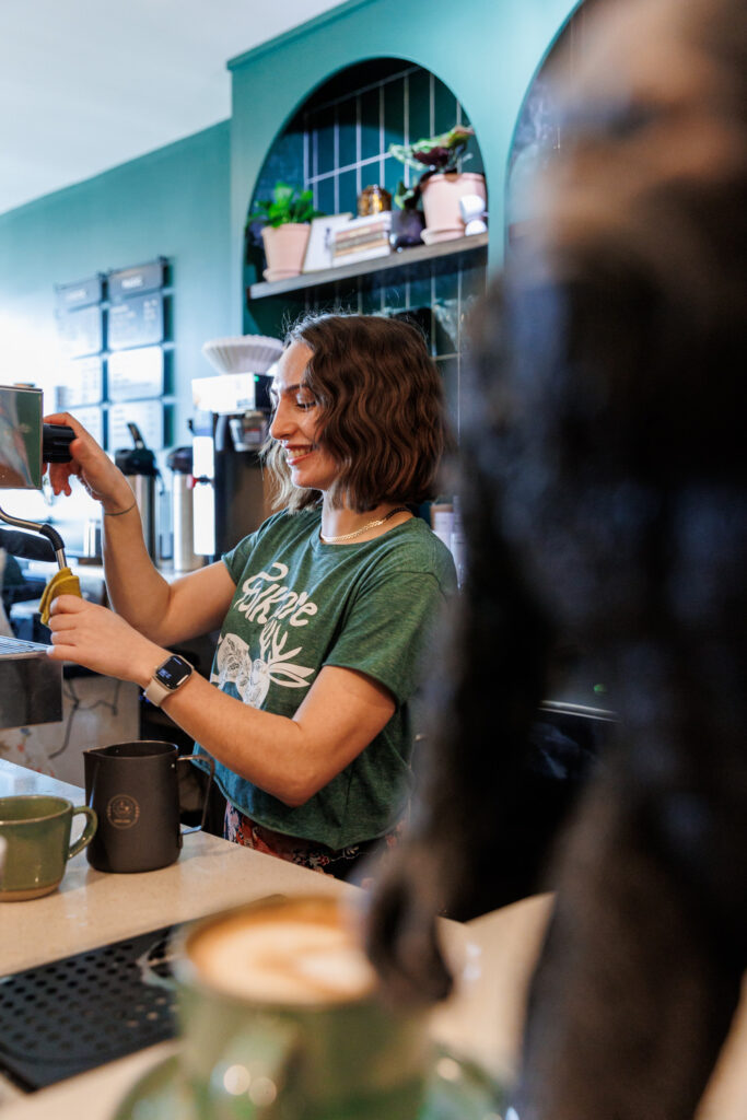 A barista prepares a coffee drink using an espresso machine in a cozy coffee shop. She is wearing a green "Folklore Coffee" T-shirt and a smartwatch, smiling as she works. The background features green-tiled shelves with potted plants and coffee supplies, contributing to the welcoming atmosphere.