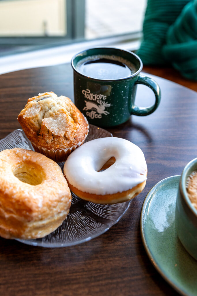 A plate with a variety of pastries, including a glazed donut, a sugar-topped muffin, and another pastry, sits on a wooden table next to a green "Folklore Coffee" mug filled with coffee. The background features a cozy setting with a green sweater and a window view.