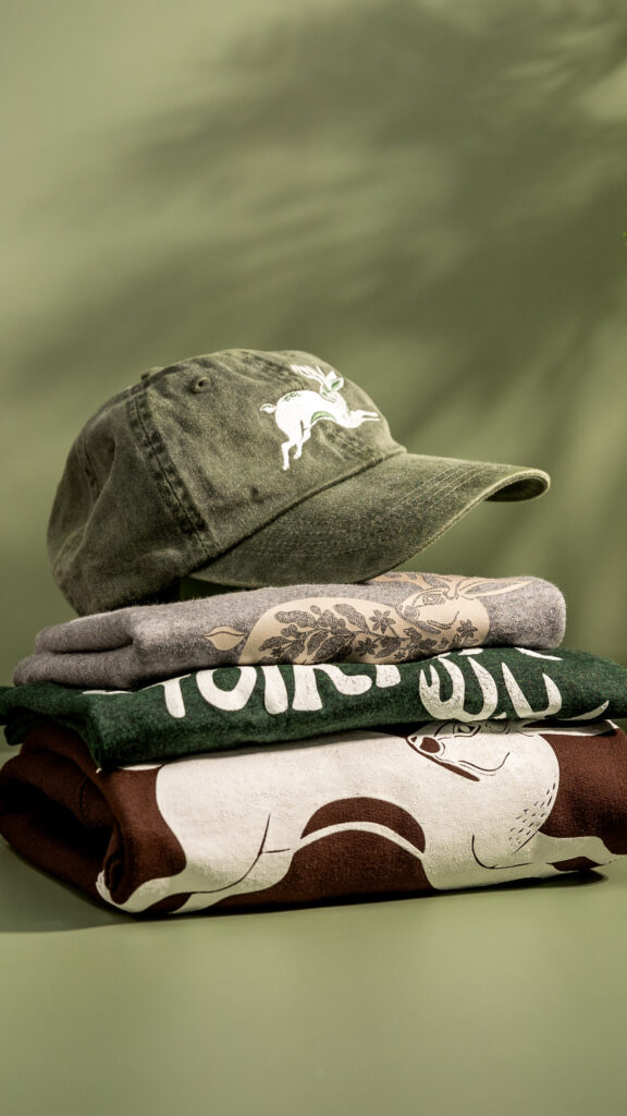 A neatly stacked arrangement of Folklore Coffee merchandise, including a green baseball cap with a white mythical creature logo on top of folded graphic T-shirts in shades of gray, green, and brown. The items are set against a soft green background, highlighting the brand's earthy and rustic aesthetic.