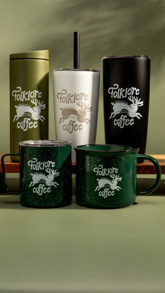 A variety of "Folklore Coffee" branded drinkware, including two green speckled mugs, an olive green tumbler, a white tumbler with a straw, and a black tumbler, are displayed against a green background. Each item features the Folklore Coffee logo with a mythical creature design.