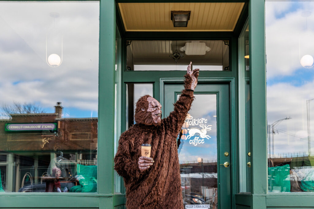 A person in a Bigfoot costume stands in front of the Folklore Coffee shop, holding a to-go cup with the Folklore Coffee logo. They are pointing upwards with their other hand, creating a playful and whimsical scene. The shop's green exterior and logo are visible, along with reflections of the surrounding buildings and sky in the glass windows.