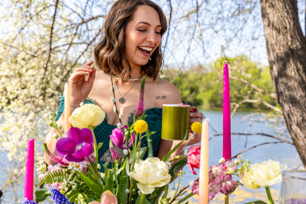 A woman joyfully holds a green mug adorned with pink flowers at an outdoor table decorated with colorful candles and vibrant spring flowers. She is dressed in a green top. The background features a scenic view of a lake and blooming trees.