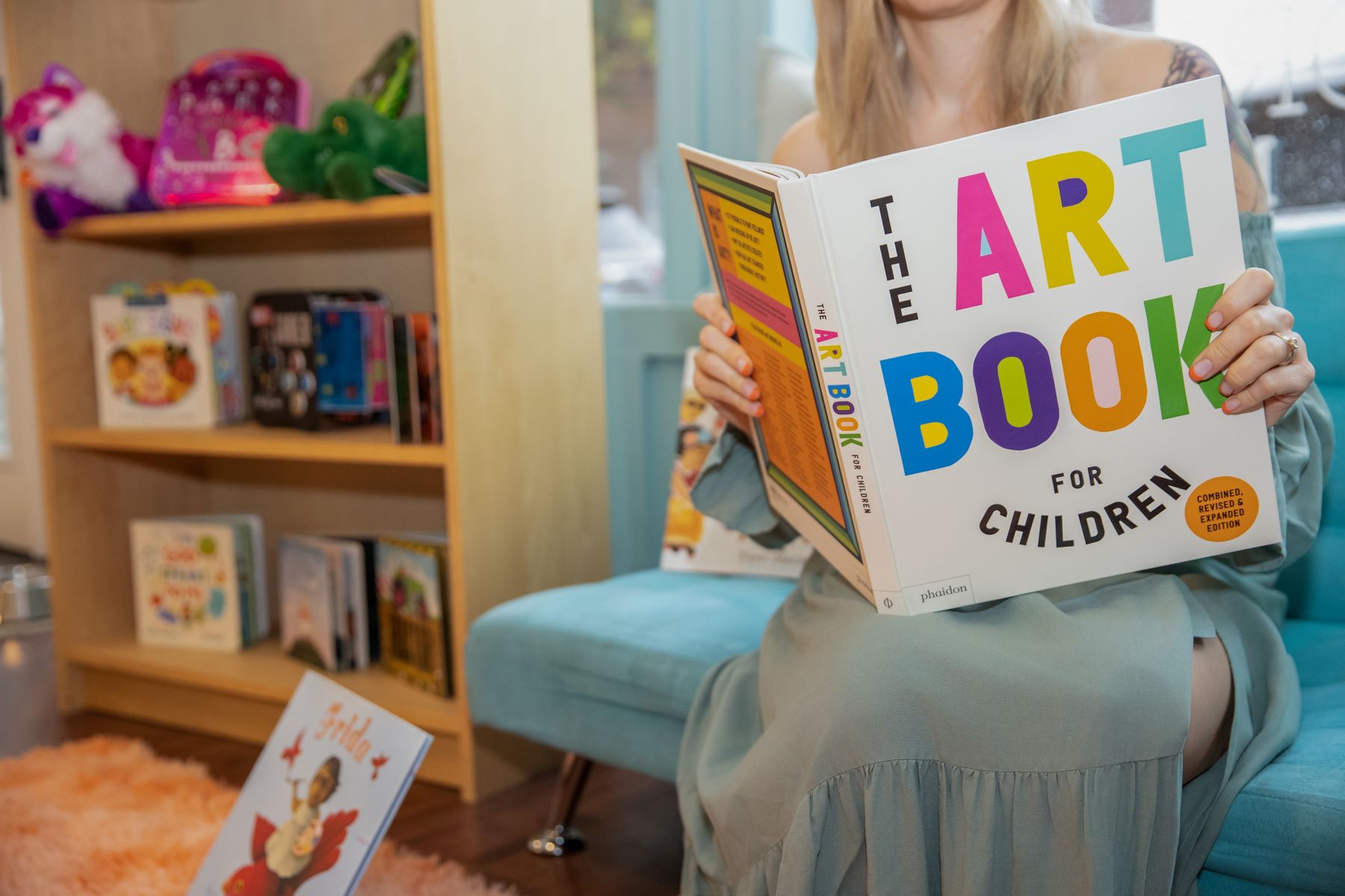 Woman sitting on a blue couch and holding a book in her hands behind her is a window and a wooden bookshelf with different books displayed 