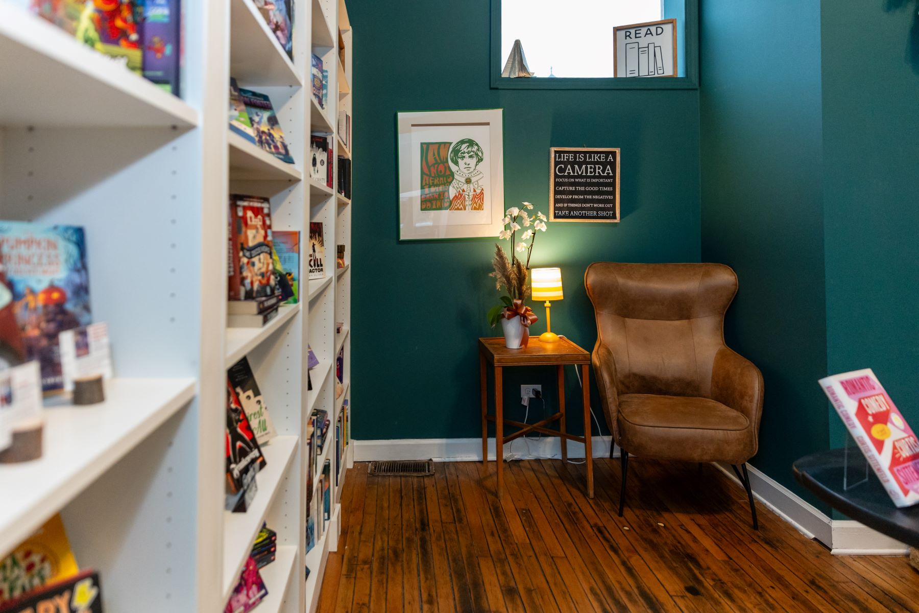 A leather chair sits in the corner next to a wooden table with a lamp and flowers and on the wall are different signs and next to the chair is a large white bookshelf with different books