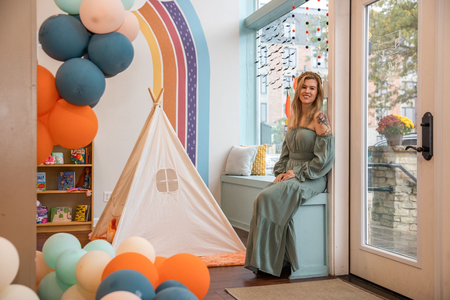 A woman is sitting next to a bay window and is surrounded by blue and orange balloons and a children's play tent with an orange rug underneath of it. The woman is wearing a mint green dress.