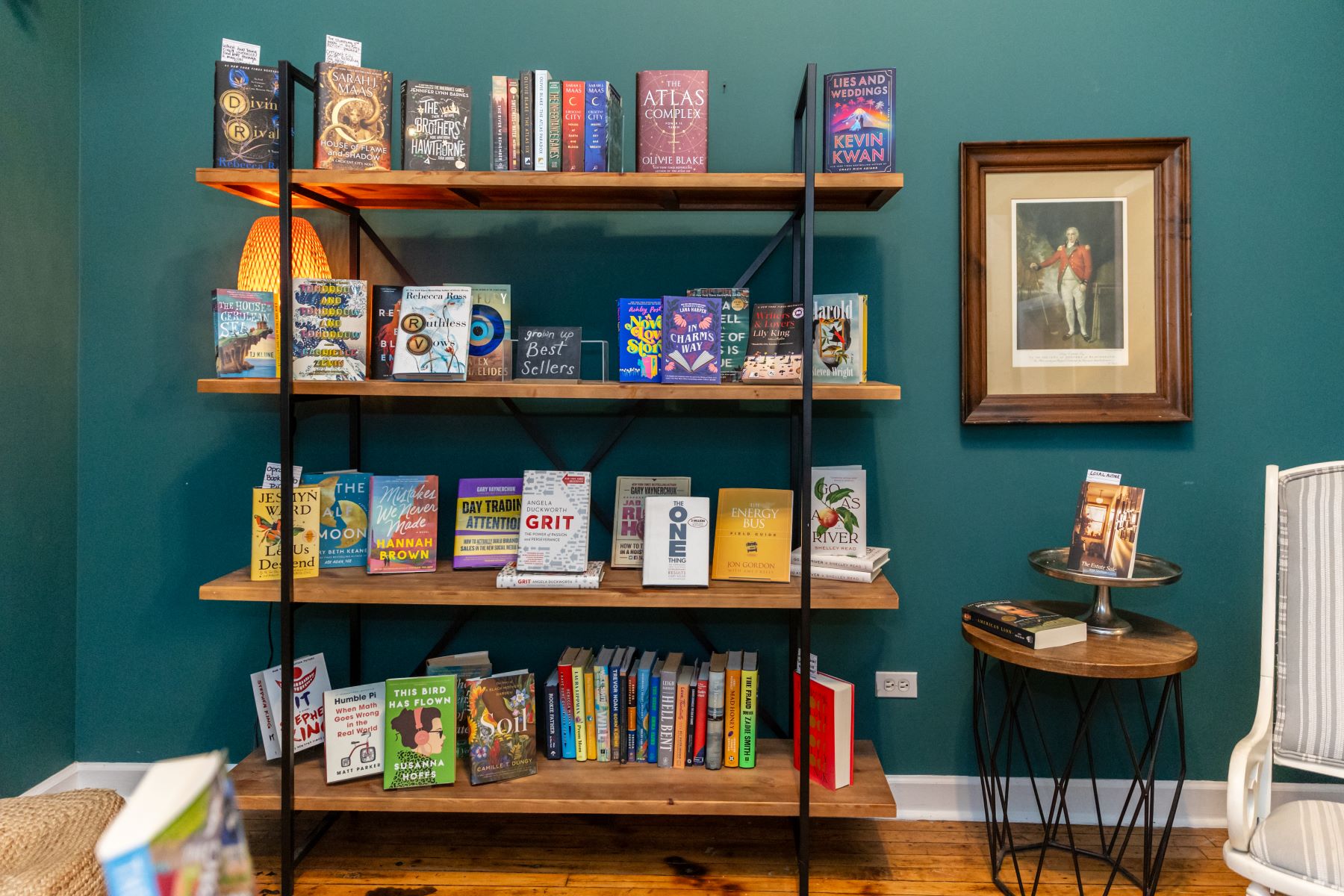 A bookshelf with a variety of different books against a green wall and there is a painting on the wall, a small side table with more books, and a striped gray chair in the corner 