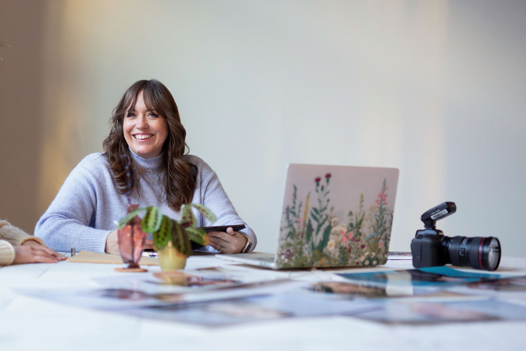 A woman in a blue sweater sitting at a table with her camera, laptop, a tablet, and with small plants and different images laid out in front of her