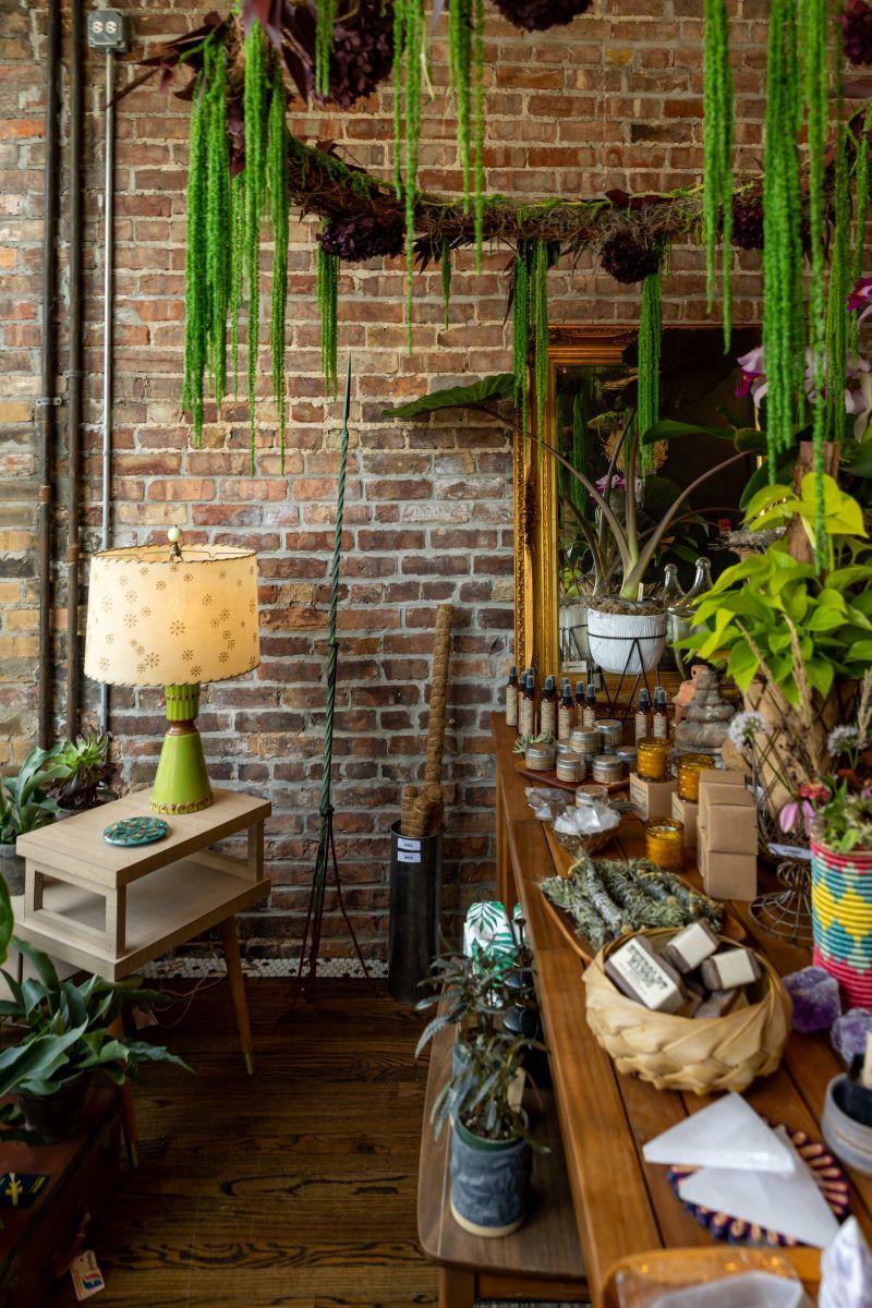 A plant store with a variety of different plants on the shelves, the ground, and hanging from the ceiling and on teh table there is also sage, candles, soap, and sprays and there is a lamp that is turned on in the corner on a wooden table there is also a brick wall in the background 