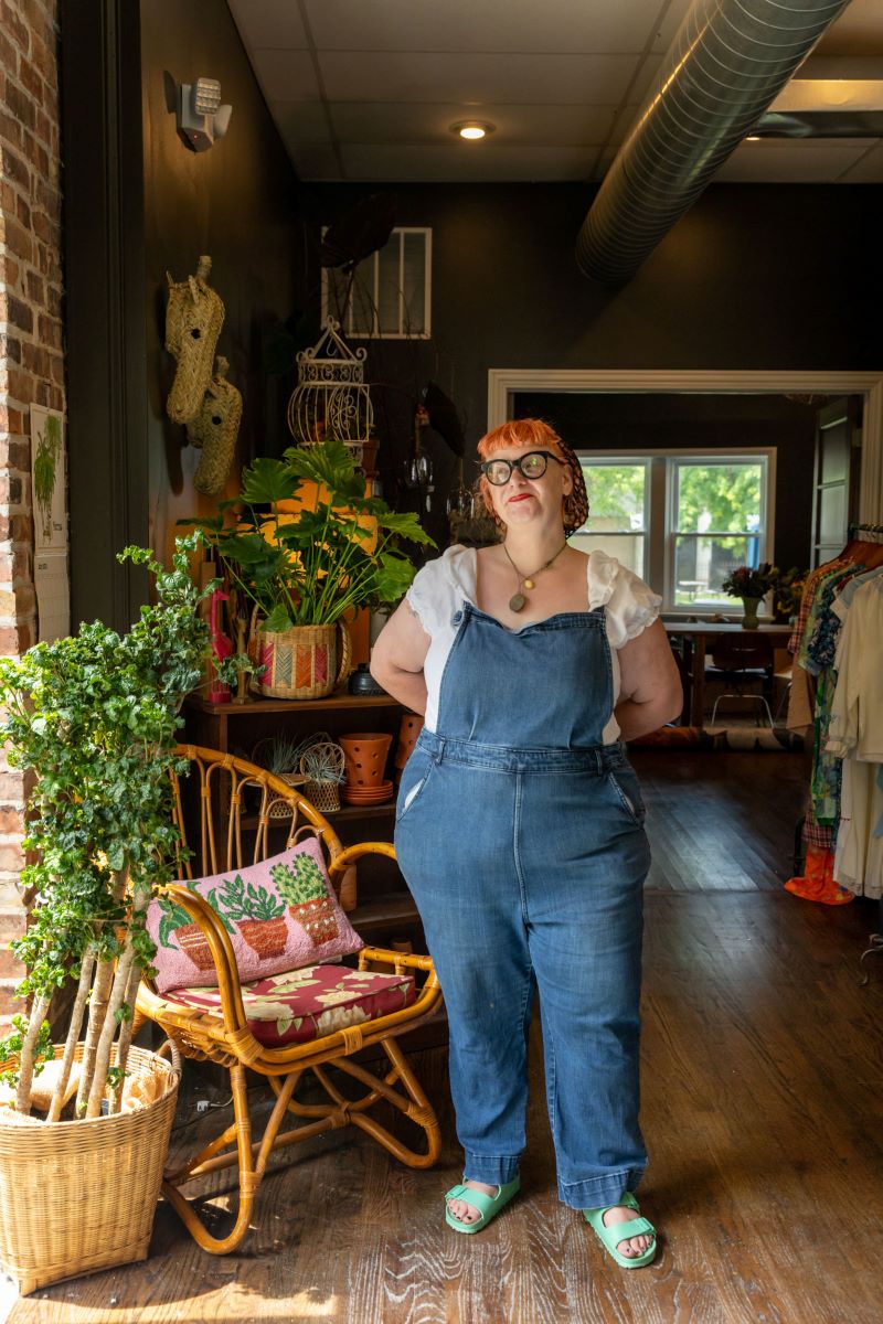 A woman in blue overalls, a white blouse, with red hair, black glasses, a necklack is standing with her arms behind her and behind the woman is a wooden wicker chair next to a brick wall and a plant in a wooden basket and also different plants and different clothes for sale inside the plant shop