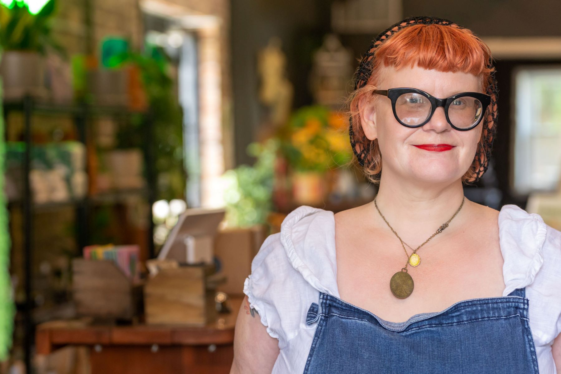 A woman with red hair, a black hair net, black glasses, red lipstick and a white top and blue overalls and wearing a necklace is softly smiling in the background is a plant shop 