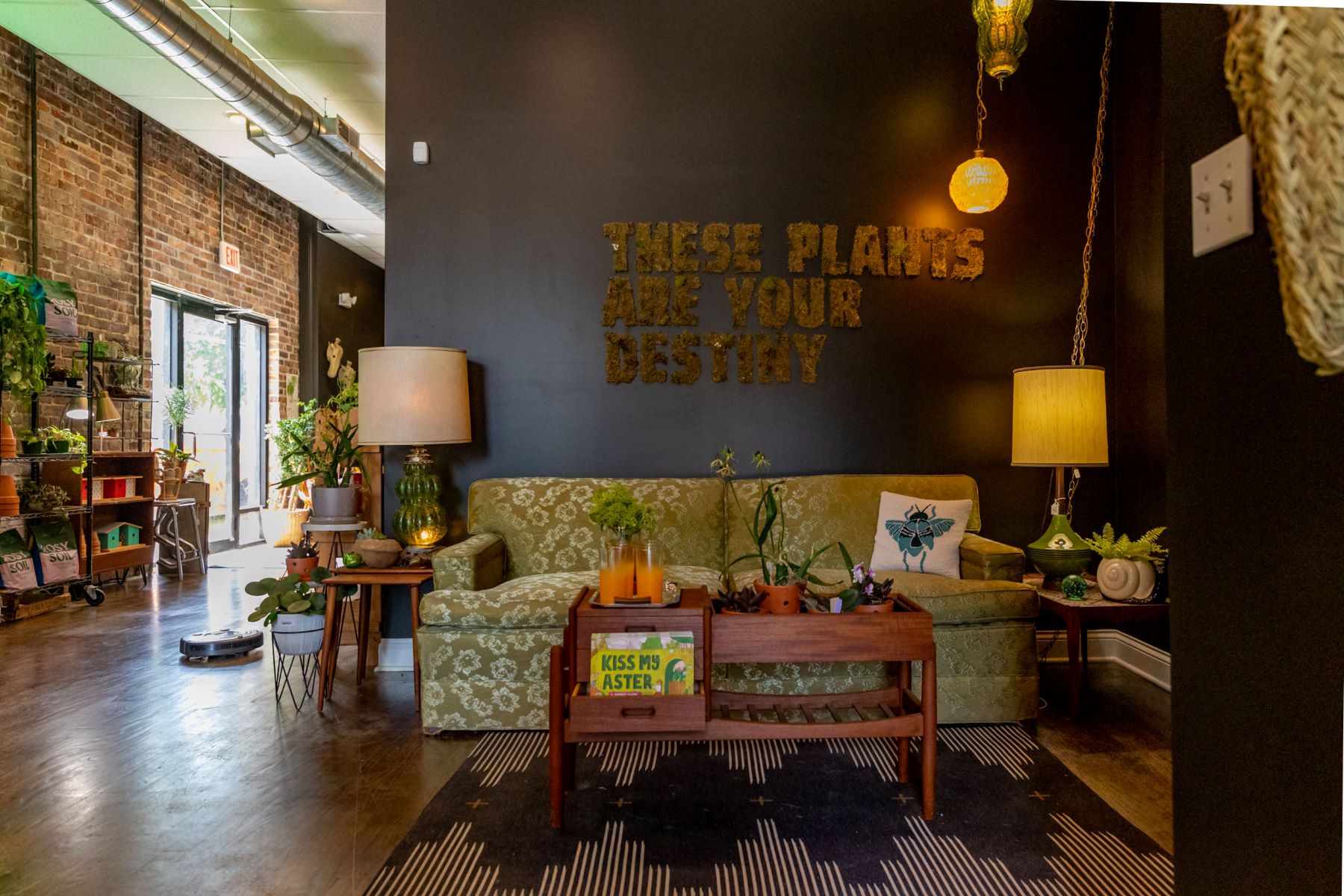 A room with a green couch with white flowers, lots of different green plants, a wooden coffee table a black rug with a white striped pattern, and lots of different lamps that are turned on there are plants on all of the different side tables a on one side is a brick wall with two different shelves of plants and a glass door 