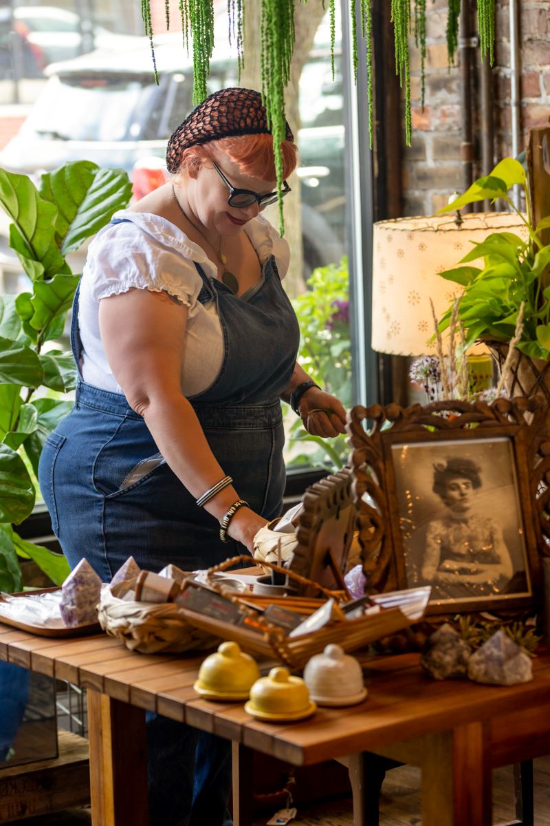 A woman in a plant shop she is wearing overalls and a white shirt, glasses, and a black hairnet she is surrounded by a variety of plants and in front of her is a wooden table with different knick knacks such as a wooden photo frame with a black and white picture, bells, and crystals 