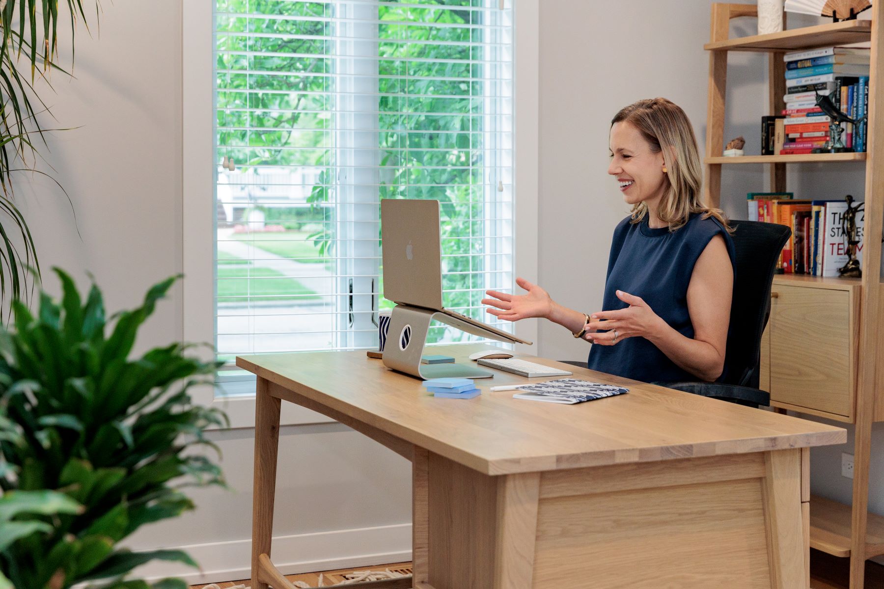 A woman in a blue shirt sitting at a desk with her laptop smiling she is in her home office space which has a large window, a bookshelf, and different plants on her desk is a coffee mug, notebook, sticky notes, and a keyboard