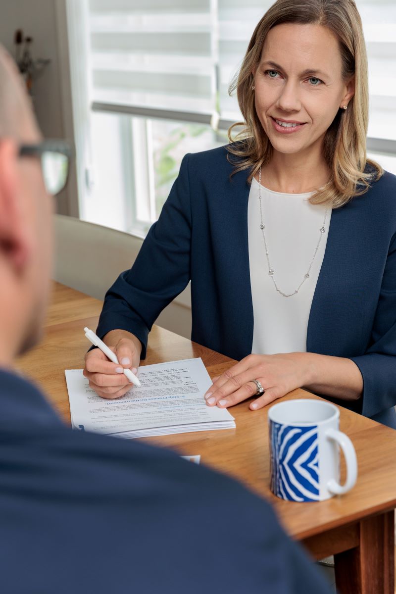 A woman in a blue blazer and white shirt is holding a pen and listening to a man in front of her and on her desk is a coffee mug and a stack of papers 