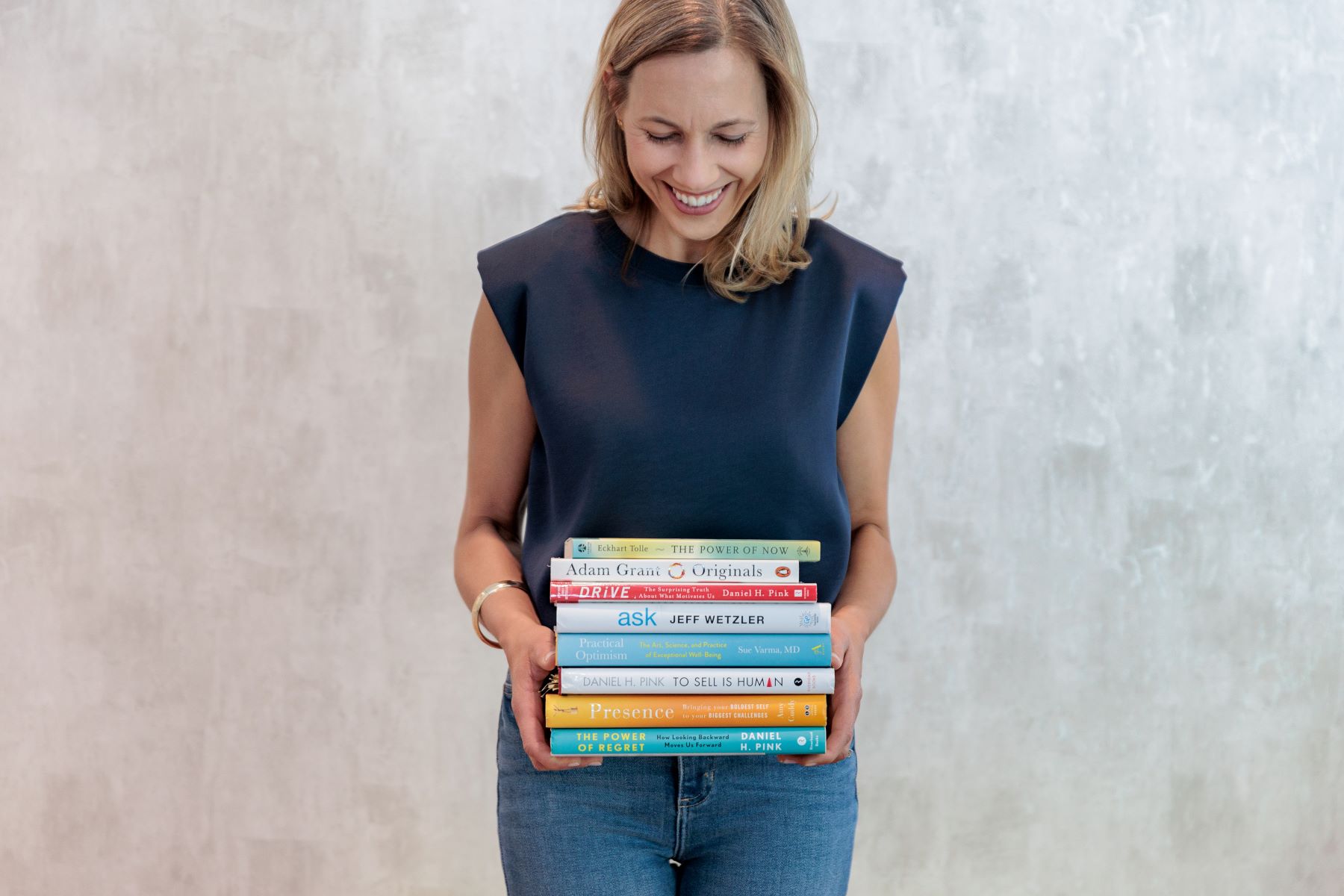 A woman in a blue shirt and jeans is holding a stack of books in her hands and smiling