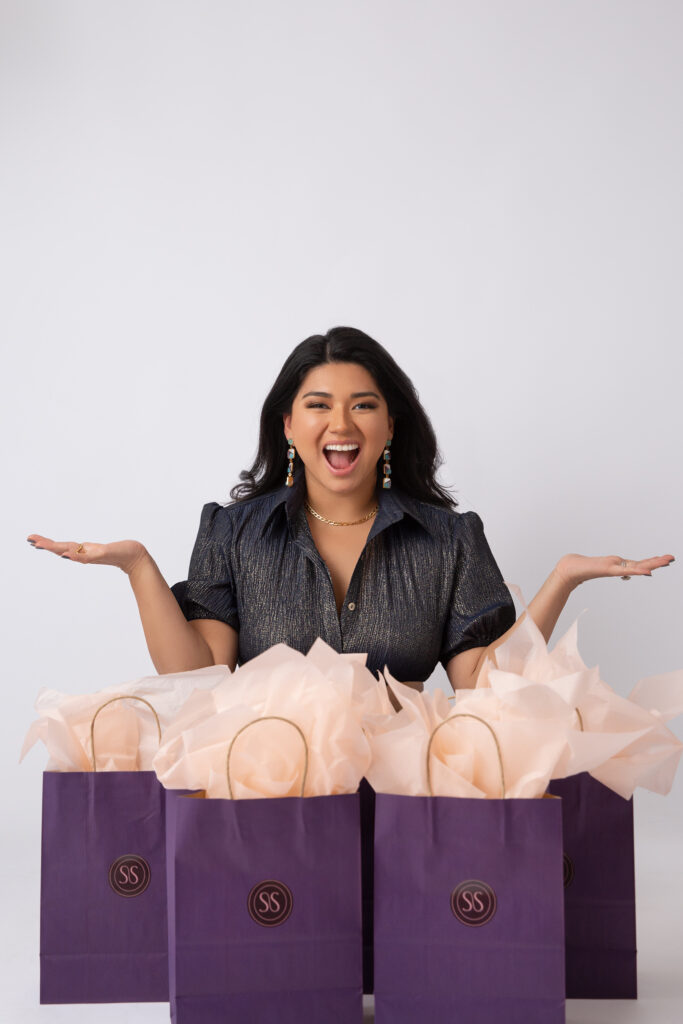 A person smiling with her hands out to either side and purple gift bags in front of her. 