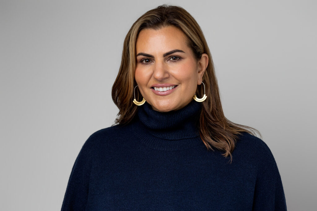 A woman smiles in a navy turtleneck and gold jewelry.