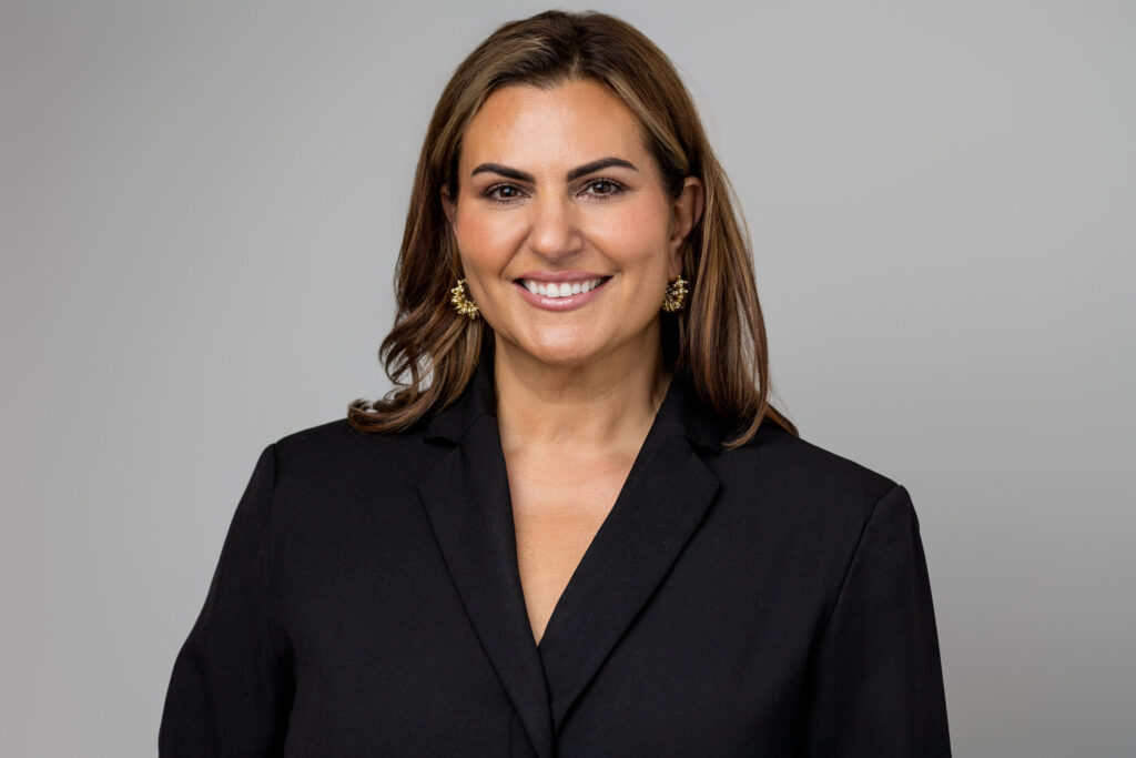 A person smiles in a black blazer and gold jewelry.