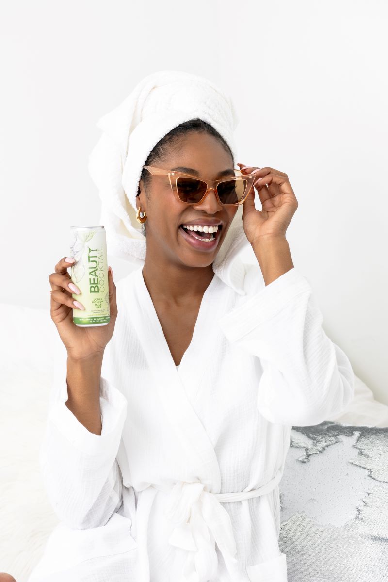 A woman sitting on a bed she is wearing sunglasses and holding a drink in one hand she is also wearing a white bathrobe and a white towel on her head