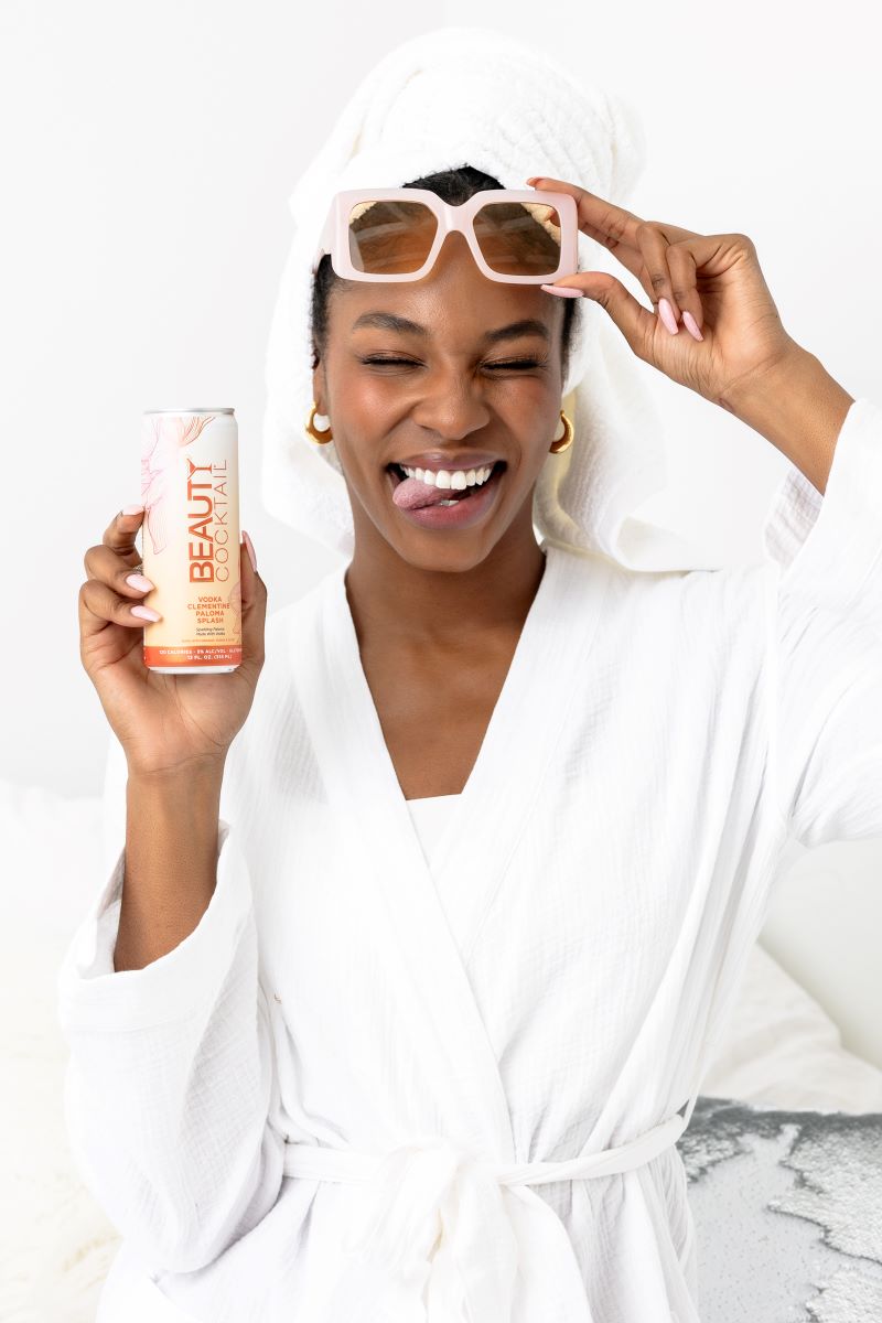 A woman wearing a towel on her head and placing sunglasses onto her head with one hand with the other she is holding and showing off a drink 