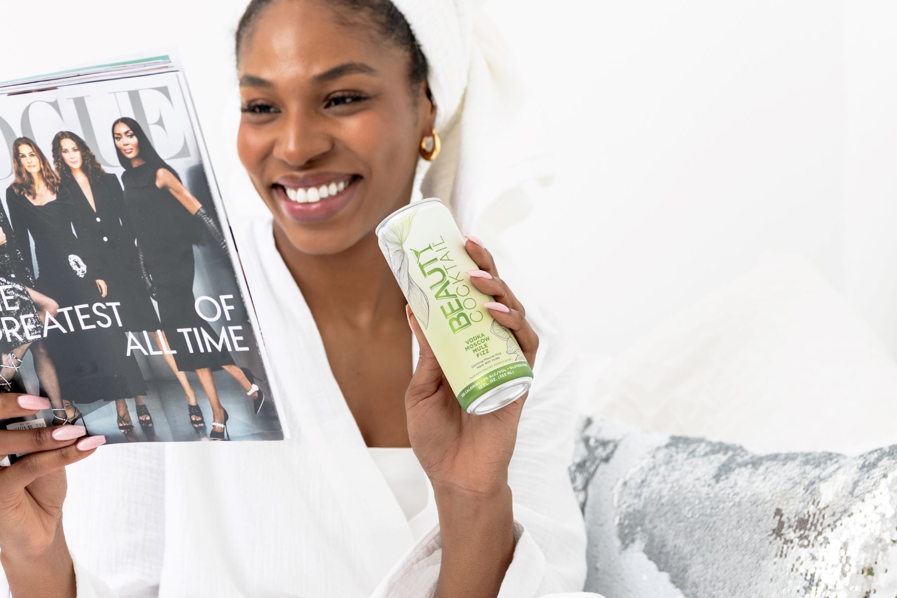 A woman in a white bathrobe with a white towel wrapped around her head is sitting on a bed and smiling and looking at a magazine and holding a drink with her other hand