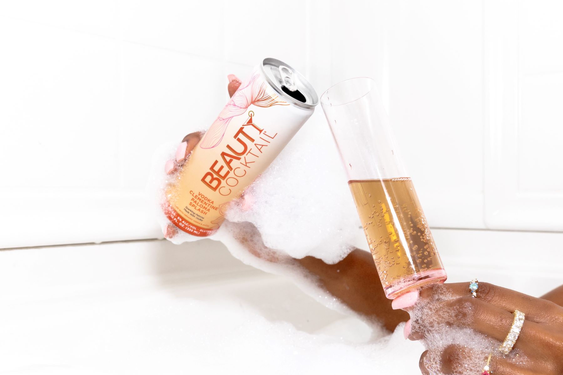 A woman sitting in a bathtub with bubbles pouring a drink into a glass 