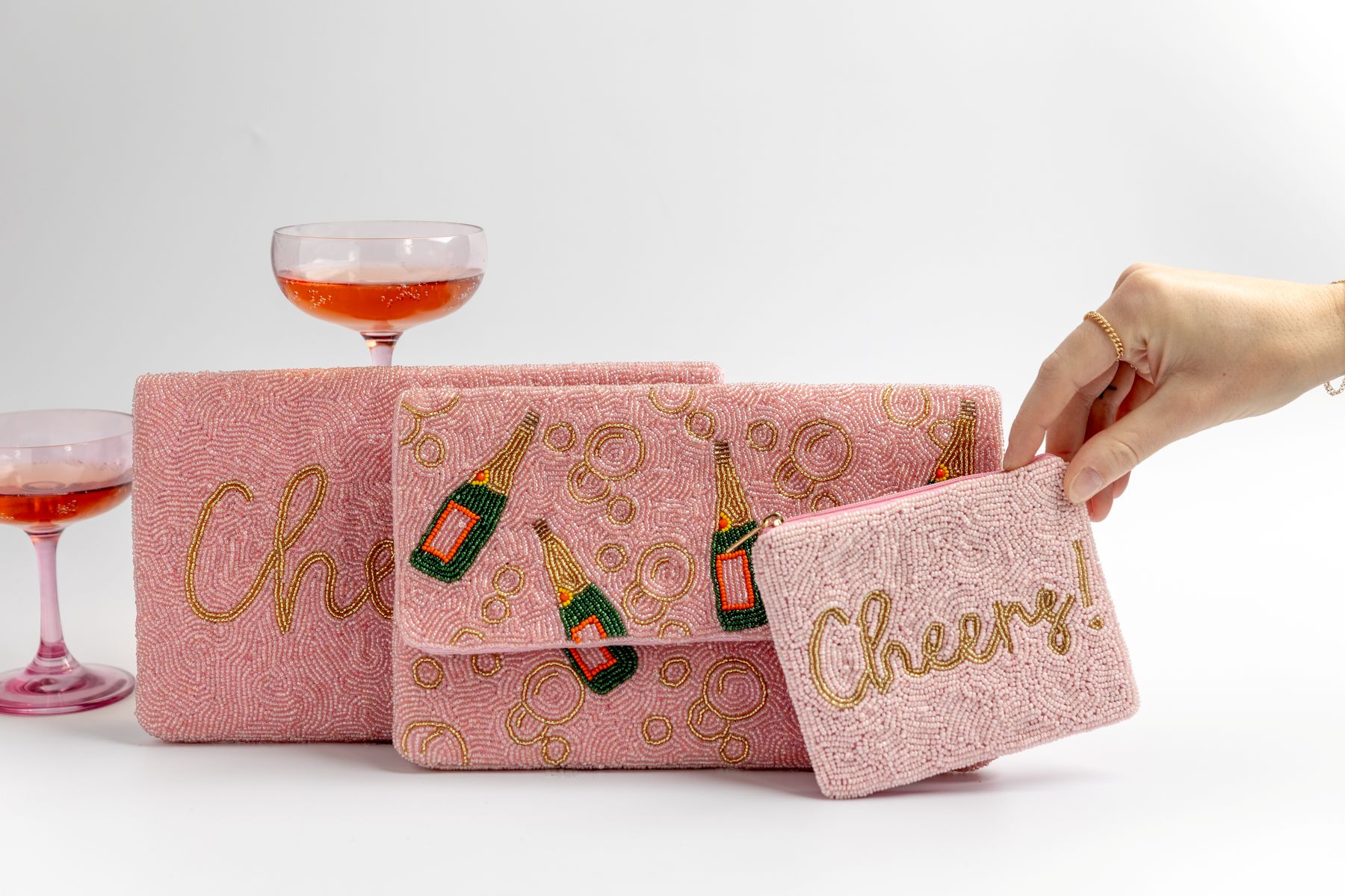 three pink wallets of varying sizes with two glasses with a drink posed behind them a hand is reaching for the smallest wallet 