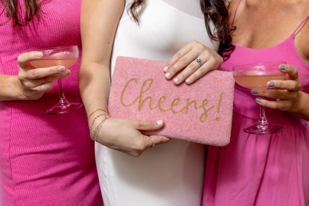 three women standing together in pink dresses one is holding a pink wallet that says cheers the other two are holding glasses with a drink in them 