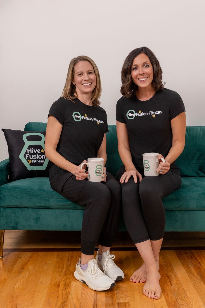two women wearing hive fusion fitness shirts sitting on a green couch and holding coffee mugs 